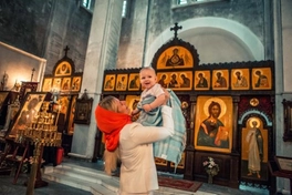 Малыш на руках у мамы в церкви, улыбается во время крещения, создавая трогательную атмосферу священного момента.